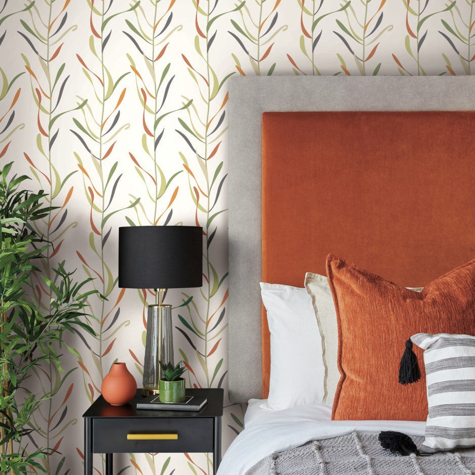 A cozy bedroom corner featuring an orange upholstered headboard, multicolored York Wallcoverings Chloe Vine River Rock Wallpaper Beige, Grey (60 Sq.Ft.), a black bedside table with a lamp, and decorative pillows. A green potted plant adds a touch of nature.