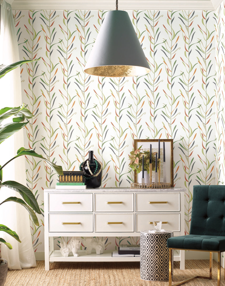 A room with whimsical design features York Wallcoverings Chloe Vine River Rock Wallpaper Beige, Grey (60 Sq.Ft.) and a white dresser with gold handles. On the dresser, there are green beaded necklaces, stacked books, candles, and framed artwork. A dark green chair sits on the right, while plants with broad leaves are on the left. A grey pendant light hangs above.