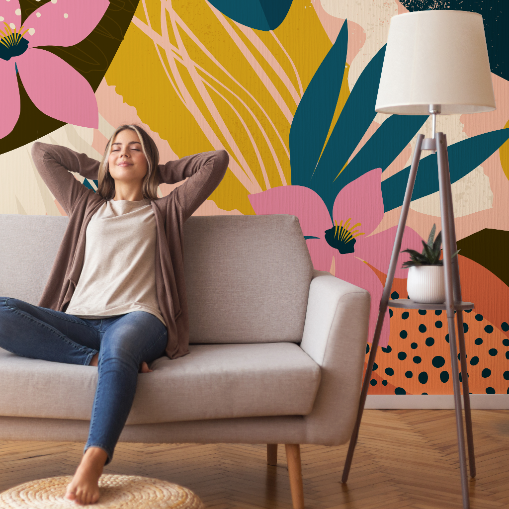 A young woman relaxes on a sofa with her eyes closed and hands behind her head in a vibrant, colorful living room with a Collage Contemporary Floral Wallpaper Mural by Decor2Go Wallpaper Mural and a floor lamp.