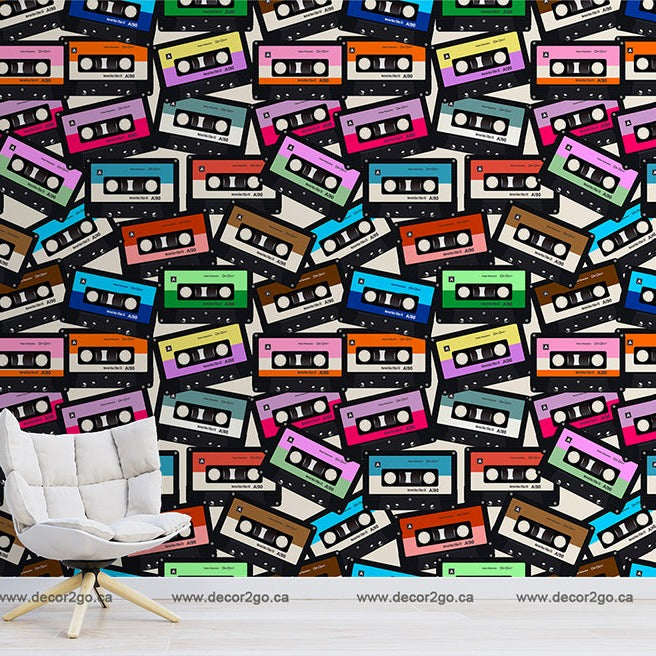 A room with a white chair against a wall covered in Decor2Go Wallpaper Mural's Coloured Cassette Wallpaper Mural of multi-colored cassette tape patterns. The floor is plain and there's a small black side table to the right of the.