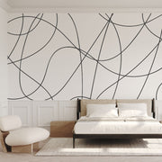 Bedroom - A minimalist bedroom featuring a large bed with white bedding and a wooden frame. The wall is adorned with the Decor2Go Wallpaper Mural's Contemporary Scribble Elegance, showcasing an artistic geometric pattern of black curved lines on a white background. To the left is a round chair, and natural light streams in through a large window with sheer curtains.
