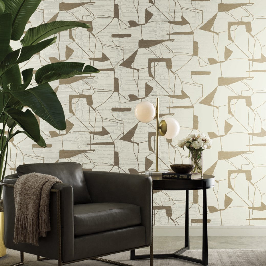 A contemporary living room corner showcases a dark leather armchair with a throw blanket, vintage gold accents including a round side table with a lamp and flowers, enhanced by lush plants. The wall is adorned with York Wallcoverings' Warm Grey & Gold Harlowe Wallpaper featuring beige and brown geometric metallic designs.