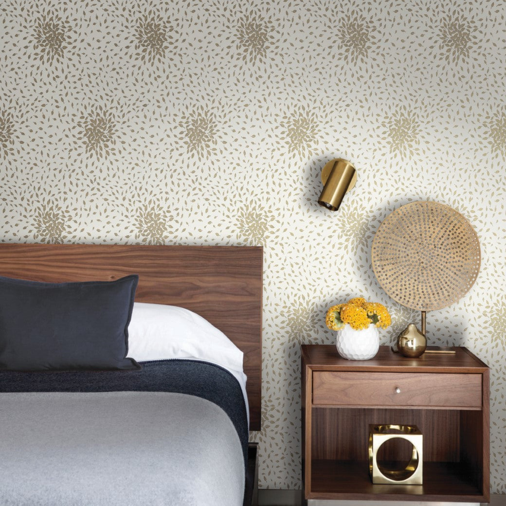 A modern bedroom features a wooden headboard, gray and white bedding, and a wooden nightstand. The wall is adorned with York Wallcoverings' Cream & Gold Petite Leaves Wallpaper. On the nightstand are a round lamp, a white vase with yellow flowers, and an elegant Modern Metals Collection piece.