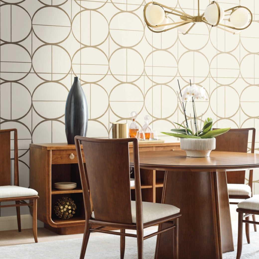 A modern dining room features a round wooden table, four chairs, a large vase, and a potted orchid. The wall displays York Wallcoverings' Cream & Gold Sun Circles Wallpaper in metallic gold circles, complemented by a contemporary chandelier above the table.