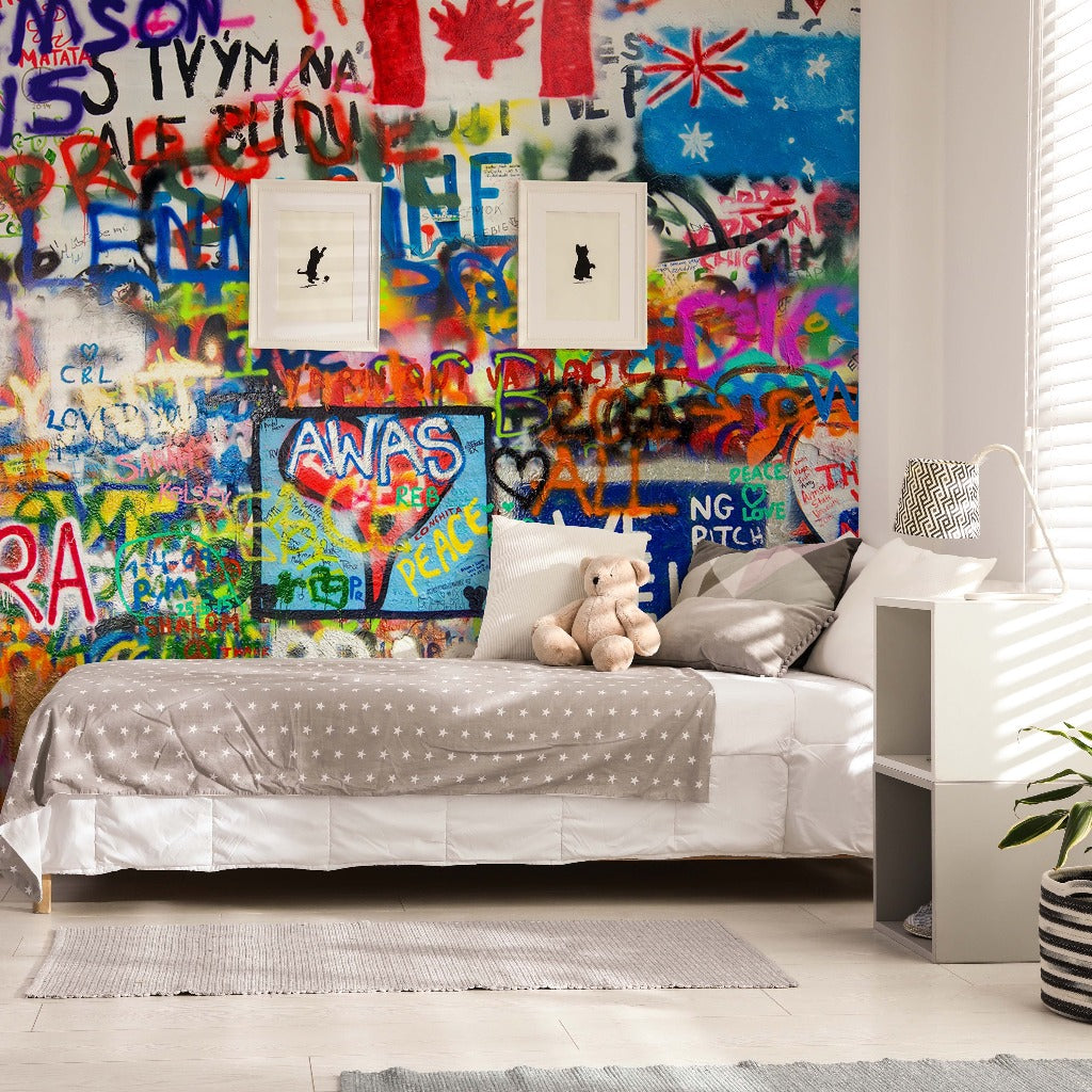 A modern bedroom featuring a single bed with a polka dot cover and a teddy bear, beside a white shelving unit. The room has vibrant, artistic colors with Decor2Go Wallpaper Mural filled with colorful graffiti design.