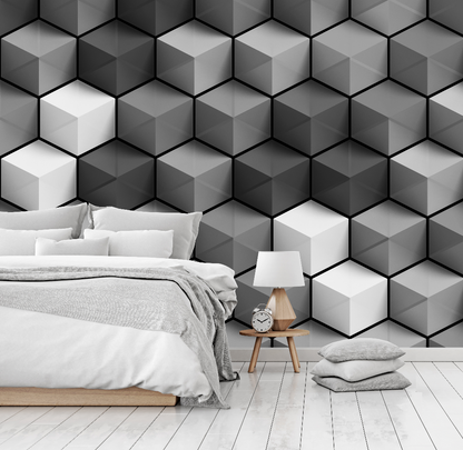 A modern bedroom features a neatly made bed with gray bedding against a Decor2Go Wallpaper Mural featuring Cubic Grays geometric wallpaper. A wooden bedside table with a lamp and clock, and two gray cushions lie on the