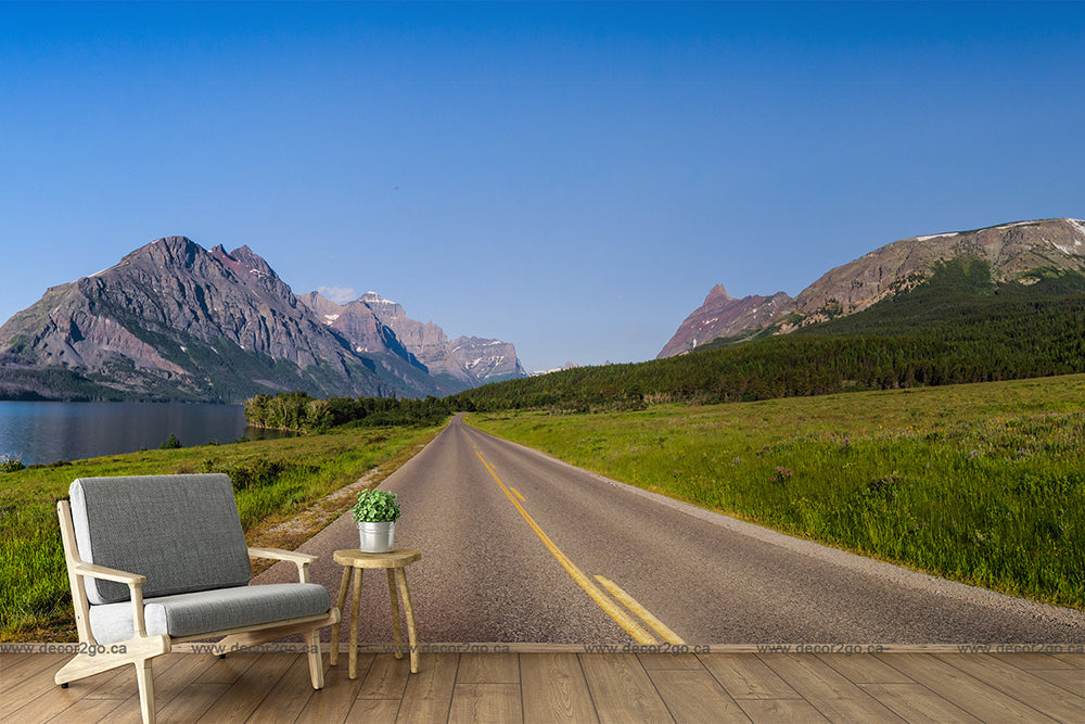 A serene landscape shows an empty road stretching toward distant mountains under a clear blue sky. To the left, a vibrant blue lake reflects the surroundings. In the foreground are a modern armchair and small table, placed on a wooden floor, like a scene from an adventurous Runaway Wallpaper Mural by Decor2Go Wallpaper Mural.