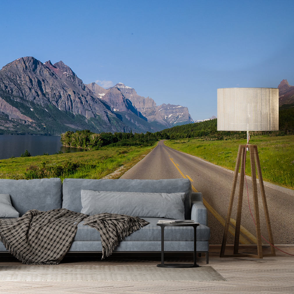 A wall mural in a modern living room features an adventurous mountain scape under a clear blue sky. The interior includes a gray couch with a checkered blanket, a small round side table, and a tall floor lamp with a beige shade decorated with the **Runaway Wallpaper Mural by Decor2Go Wallpaper Mural**.