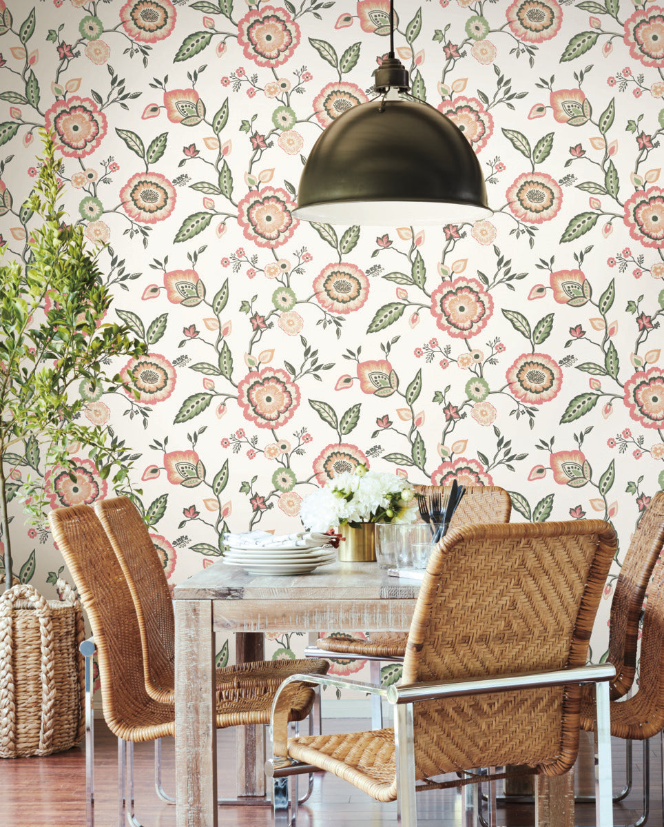 A cozy dining area features a wooden table set with dishes and flowers in a vase. The setting includes woven chairs with metal frames. The background displays botanical elegance with Dahlia Blooms Midnight/Multi Wallpaper Black, Pink (60 Sq.Ft.) by York Wallcoverings. A modern black pendant lamp hangs above the table.
