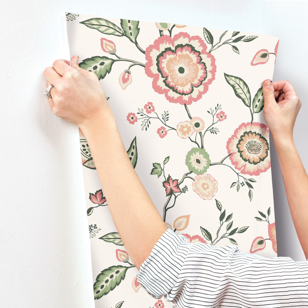 A person with a striped sleeve is applying York Wallcoverings' Dahlia Blooms Midnight/Multi Wallpaper Black, Pink (60 Sq.Ft.) to a light-colored wall. The wallpaper, featuring large flowers in shades of pink and peach with green leaves and stems, offers botanical elegance. The person's hands are smoothing out the paper, ensuring an easy installation and removal process.