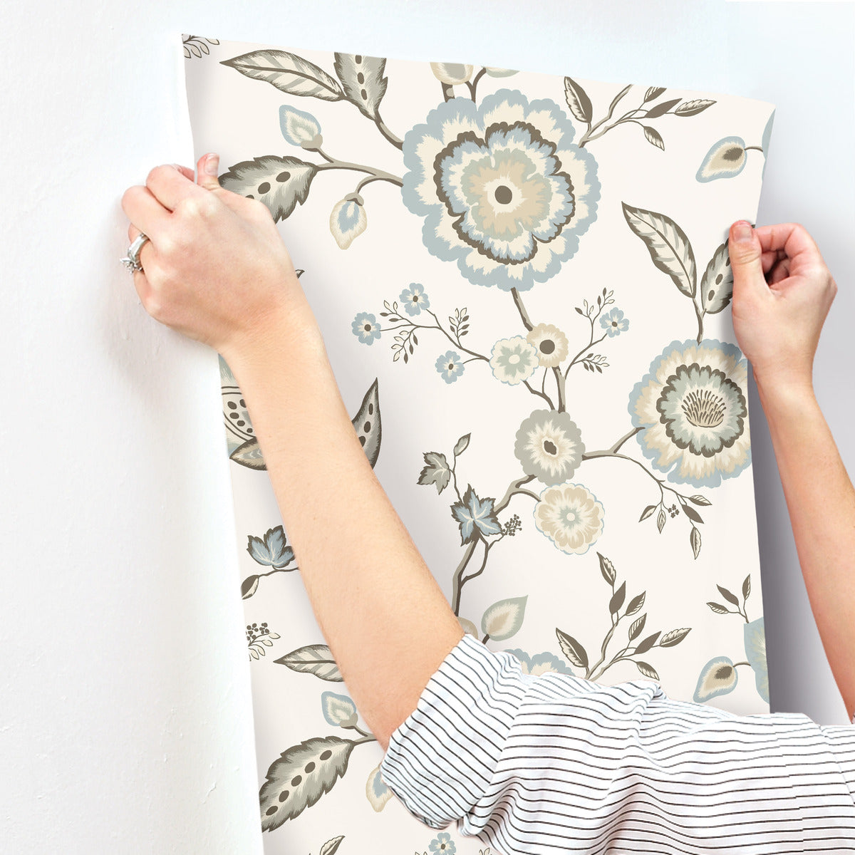 A person is holding up a sheet of *Dahlia Blooms Midnight/Multi Wallpaper Black, Pink (60 Sq.Ft.)* by York Wallcoverings with a floral design in shades of blue, gray, and beige against a blank white wall. The person wears a long-sleeved, striped shirt and has a ring on their left hand. The wallpaper features large flowers, leaves, and small buds, offering *Botanical Elegance*.