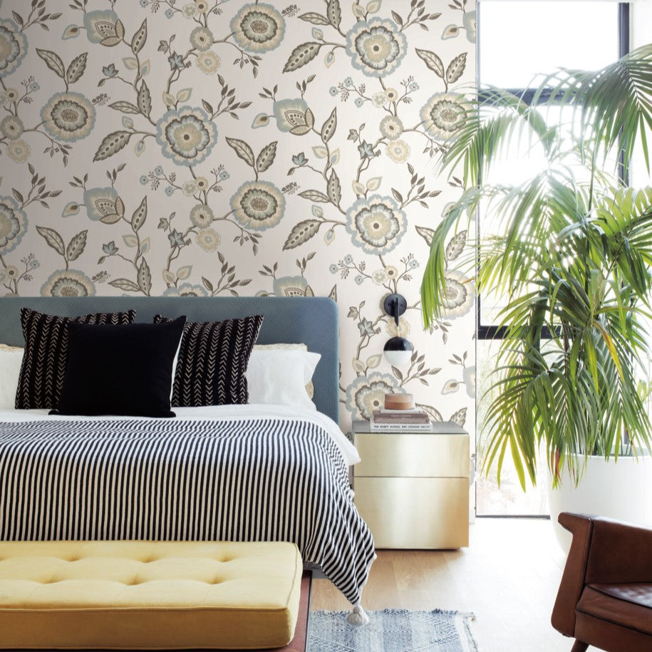 A modern bedroom features a bed with a grey upholstered headboard, white and black pillows, and a striped duvet. The wallpaper, showcasing York Wallcoverings Dahlia Blooms Dove/Cornflower Wallpaper Blue, White/Off White (60 Sq.Ft.), adds botanical elegance. A yellow bench is at the foot of the bed, and a large plant is near a window letting in natural light.