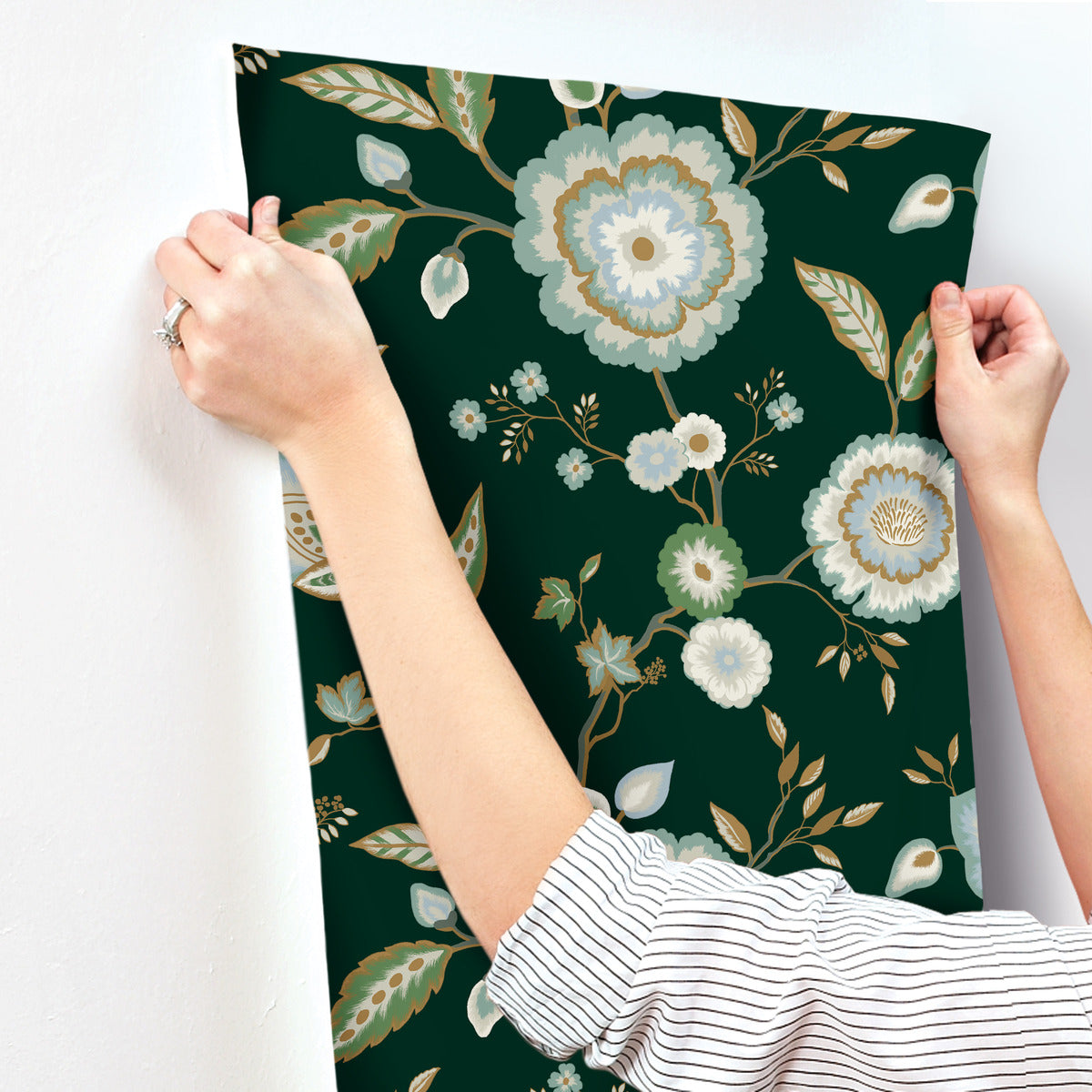 A person is applying Dahlia Blooms Midnight/Multi Wallpaper Black, Pink (60 Sq.Ft.) by York Wallcoverings. The person's hands are visible, holding the wallpaper in place, and they are wearing a striped shirt with sleeves rolled up.