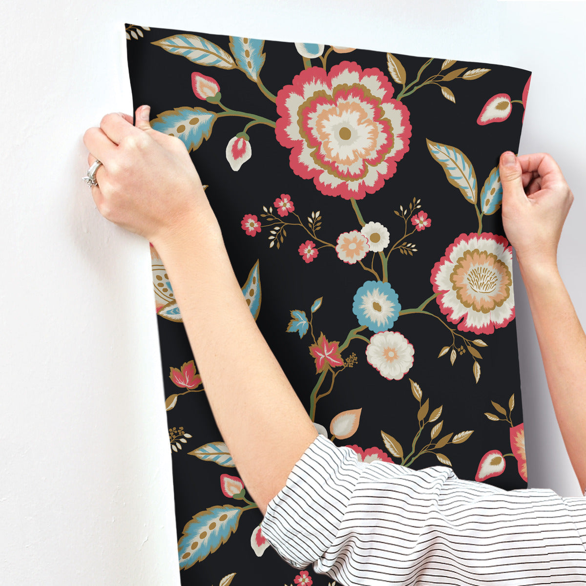 A person is hanging up a piece of York Wallcoverings' Dahlia Blooms Dove/Cornflower Wallpaper Blue, White/Off White (60 Sq.Ft) with a floral design. The wallpaper boasts a black background adorned with colorful flowers and leaves in shades of red, pink, blue, and white. Demonstrating easy installation, the person holds the wallpaper at the top corners, aligning it on a white wall.