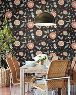 A dining area with a wooden table surrounded by wicker chairs and a metal chair. The wall is adorned with York Wallcoverings' Dahlia Blooms Dove/Cornflower Wallpaper Blue, White/Off White (60 Sq.Ft.), adding botanical elegance to the space. A hanging industrial-style lamp is above the table, which is set with white dishes, glassware, and a floral centerpiece. A potted plant is in the corner.