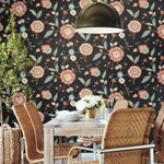 A dining area with a wooden table and six rattan chairs under a brass pendant light. The table is set with white plates and a flower vase. The wall is adorned with vibrant Dahlia Blooms Midnight/Multi Wallpaper Black, Pink (60 Sq.Ft.) by York Wallcoverings. A tall plant is in the corner, adding botanical elegance.
