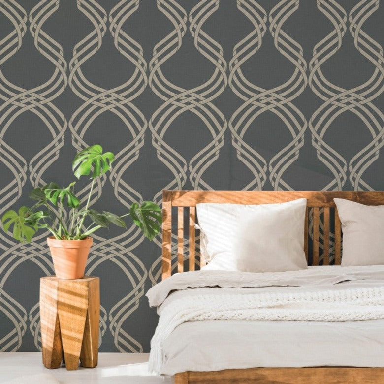 A cozy bedroom with a wooden slatted headboard bed adorned with white bedding. A potted plant with green leaves sits on a wooden stool to the left. The wall features Dante Ribbon Shaped Wallpaper (60 SqFt) by York Wallcoverings, showcasing iconic patterns in a dark geometric design that adds character to the interior décor.