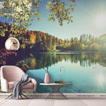 A serene lakeside scene with a pink armchair and small table, framed by lush trees and a clear blue sky, reflecting sunlight in a tranquil lake. A hanging lamp and a cozy blanket add comfort with the Day on The Lake Wallpaper Mural from Decor2Go Wallpaper Mural.