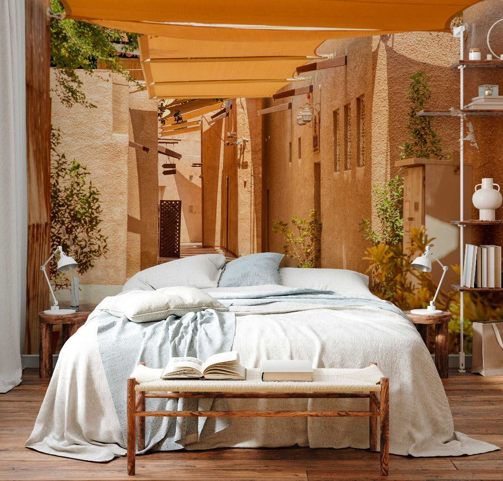 An outdoor bedroom setup in a narrow alley, featuring a large bed with blue and white bedding, wooden floor, and beige fabric shades above. A small bench with an open book is at the foot of Decor2Go Wallpaper Mural.