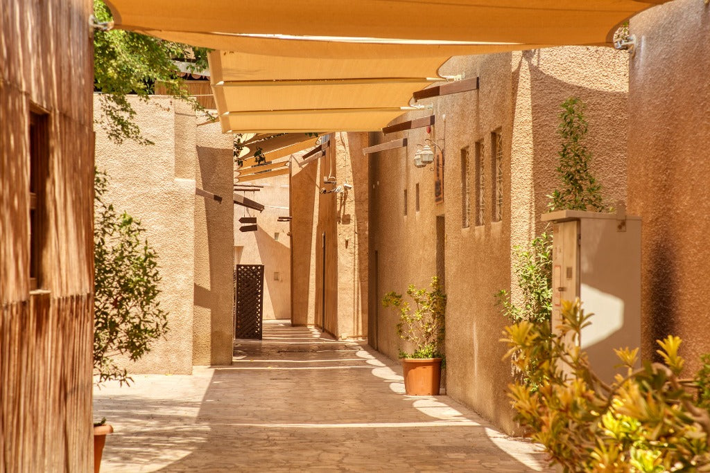 A sunny alleyway lined with beige, textured walls under a canopy, featuring potted plants and a wooden beam on the left side. This Decor2Go Wallpaper Mural conveys a warm, serene atmosphere.