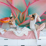 Two children having a pillow fight on a bed, with a vibrant Desert Oasis wallpaper mural in the background. one child is wearing a unicorn headband and both are smiling joyously.