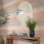 A wooden console table exudes rustic elegance, displaying a black vase with green and white flowers. Stacked books and a decorative object enhance the charm, while a large, uniquely shaped mirror on the wall above complements the scene against the Desert Textile Wallpaper in Pink by York Wallcoverings. Nearby, a potted plant breathes life into the setting.