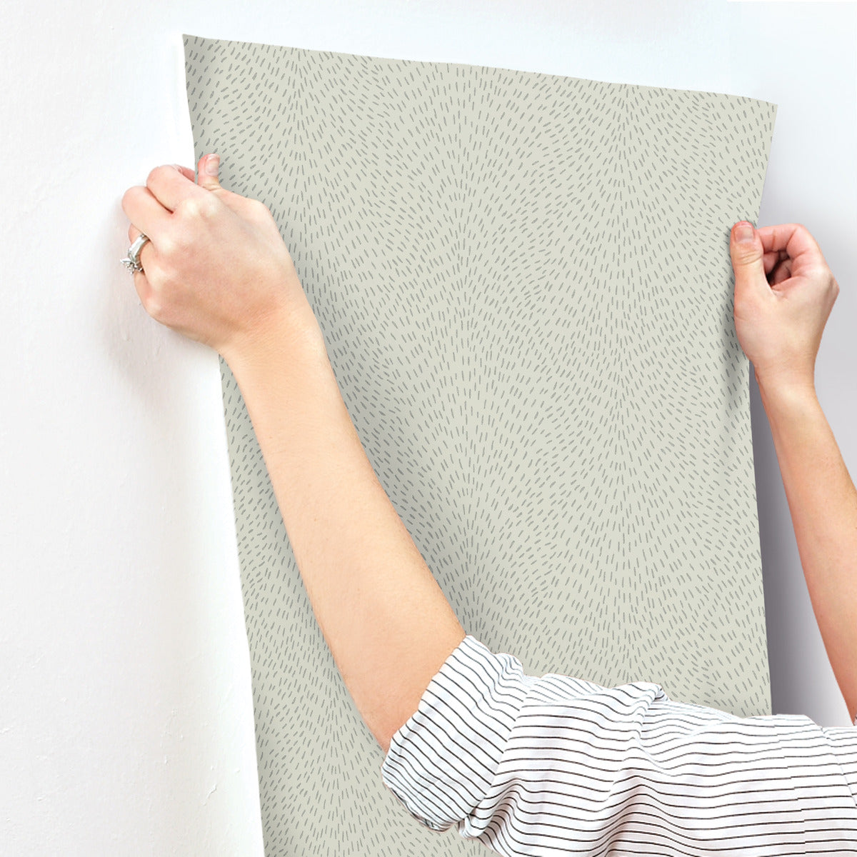 A person in a striped shirt is applying York Wallcoverings Dew Drop Grey Wallpaper (60 SqFt), featuring a light gray pattern with small, scattered dark lines, to a white wall and smoothing it with both hands.