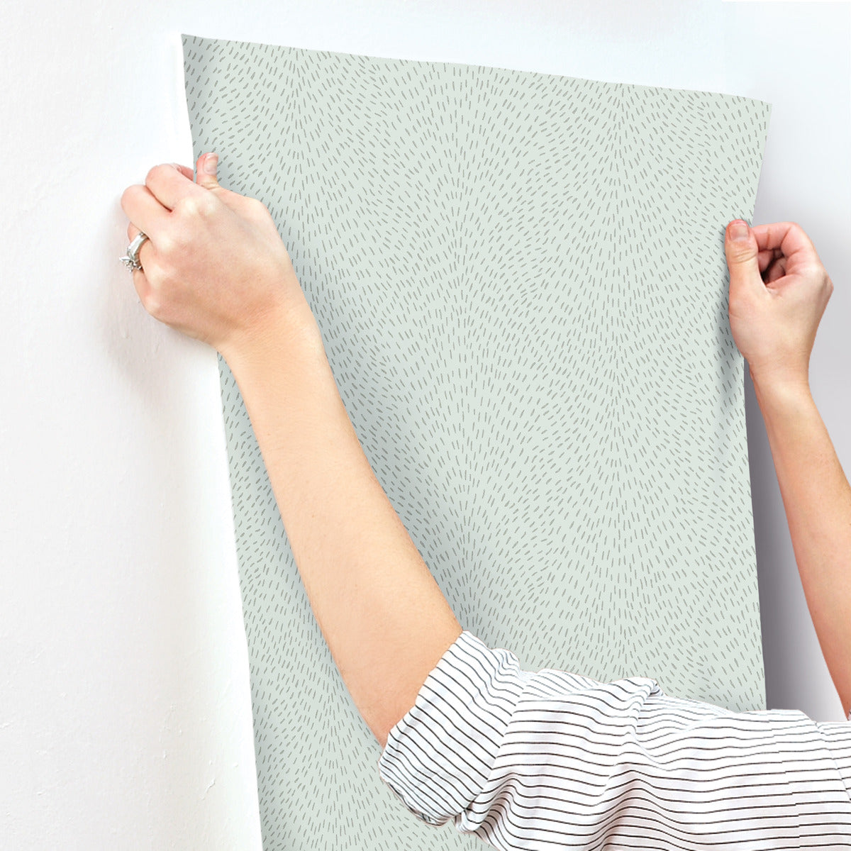 A person is carefully applying Dew Drop Sea Mist Wallpaper by York Wallcoverings to a white wall, with only their arms and hands visible as they smooth the pale blue, subtly dashed design in place.
