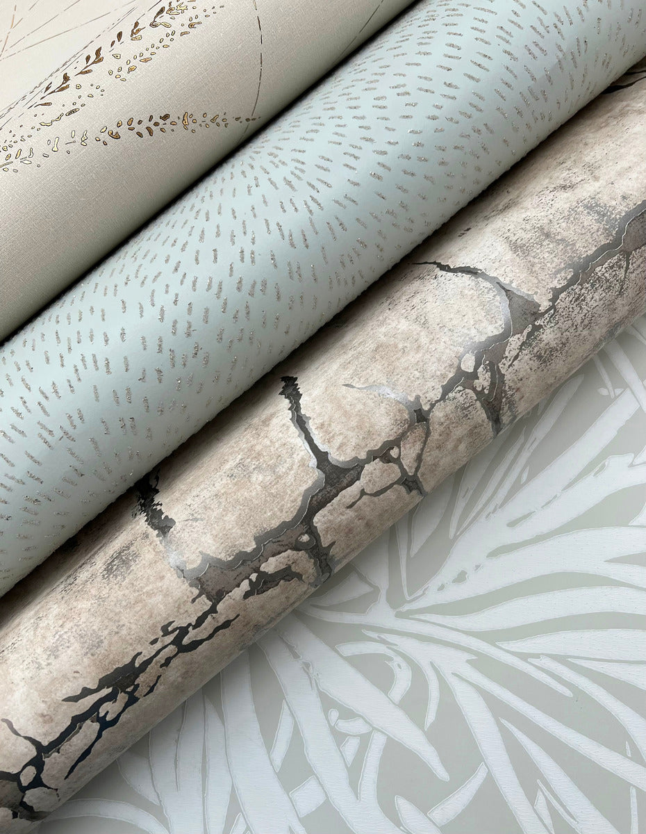 Three rolls of York Wallcoverings Dew Drop Sea Mist Wallpaper (60 SqFt) in soft neutrals are stacked, featuring gold botanicals, dotted lines, and a cracked stone look. A subtle leaf pattern accents the surface beneath the pale blue wallpaper.