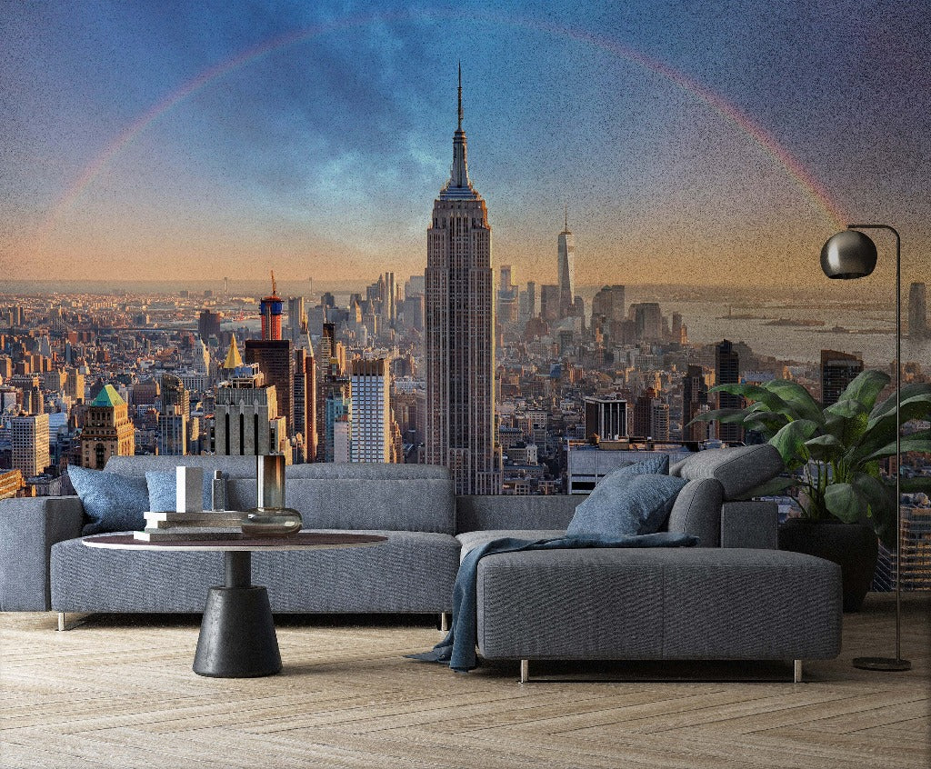 A modern living room features a Decor2Go Wallpaper Mural titled Double Rainbow Skyline with an iconic skyscraper at the center and a subtle rainbow in the sky. The room includes a sectional sofa, a round coffee table, and a floor lamp, with potted plants adding a touch of greenery to the urban aesthetic decor.