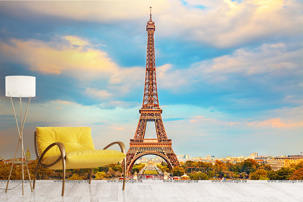 A room features a tall floor lamp with a white shade and a mustard-yellow cushioned armchair against an Eiffel Tower II Wallpaper Mural by Decor2Go Wallpaper Mural set against a colorful sunset sky. The floor is light-colored wood, and the overall decor is modern and minimalist.