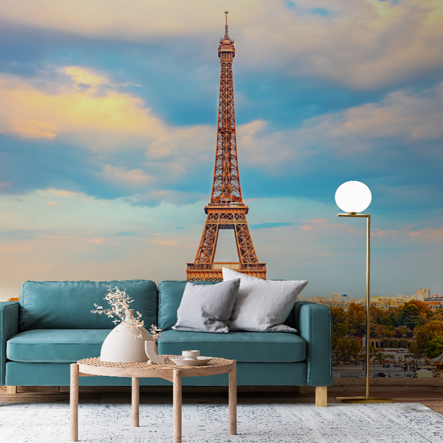 A cozy living room features a teal sofa with gray and white pillows, and a round wicker coffee table with a flower vase. A tall, modern lamp stands nearby. The backdrop is an expansive Decor2Go Wallpaper Mural's Eiffel Tower II Wallpaper Mural against a sunset sky.