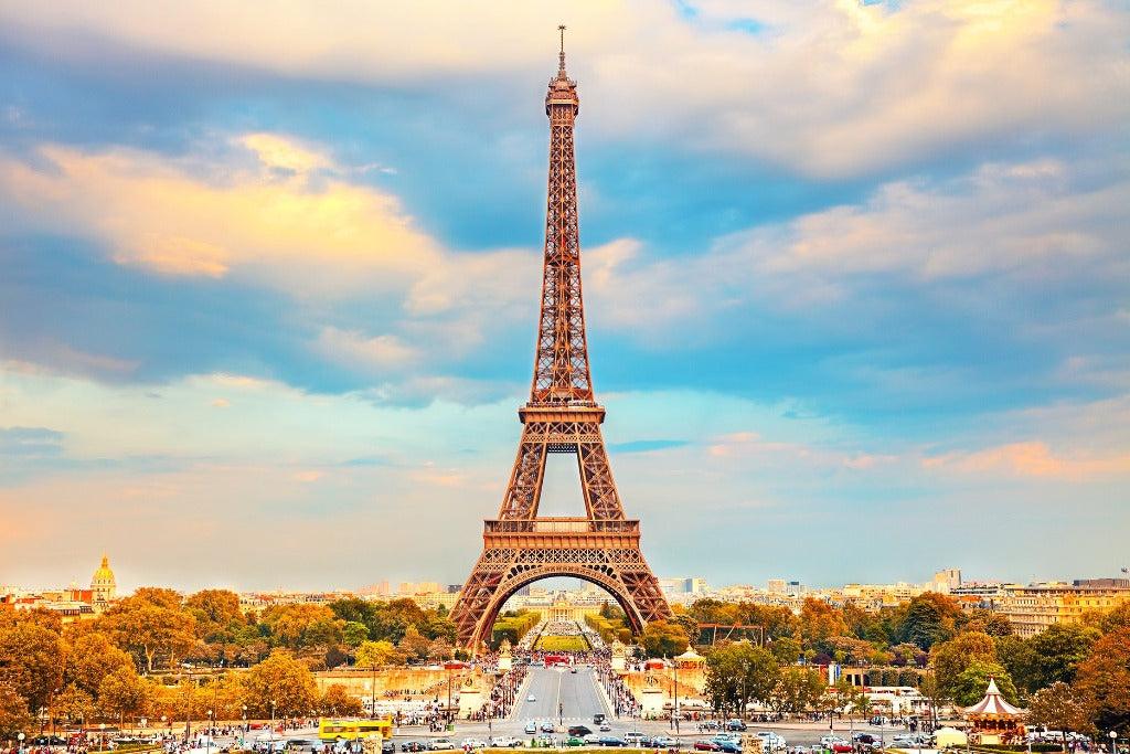 The image shows the Eiffel Tower in Paris, France, under a partly cloudy sky during sunset. The tower stands prominently in the center, with trees, buildings, and the bustling cityscape in the background bathed in the warm glow of the setting sun—perfect for an Eiffel Tower II Wallpaper Mural by Decor2Go Wallpaper Mural.