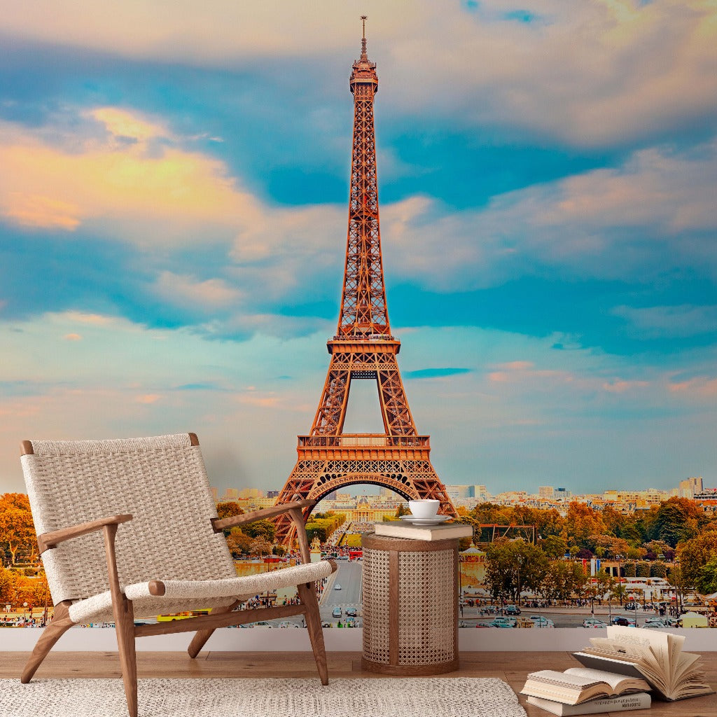 A cozy outdoor seating area with a woven chair, a small round table, and an open book. In the background, an Eiffel Tower II Wallpaper Mural by Decor2Go Wallpaper Mural rises majestically against a colorful sky at sunset. The scene combines relaxation with picturesque Paris travel decor.