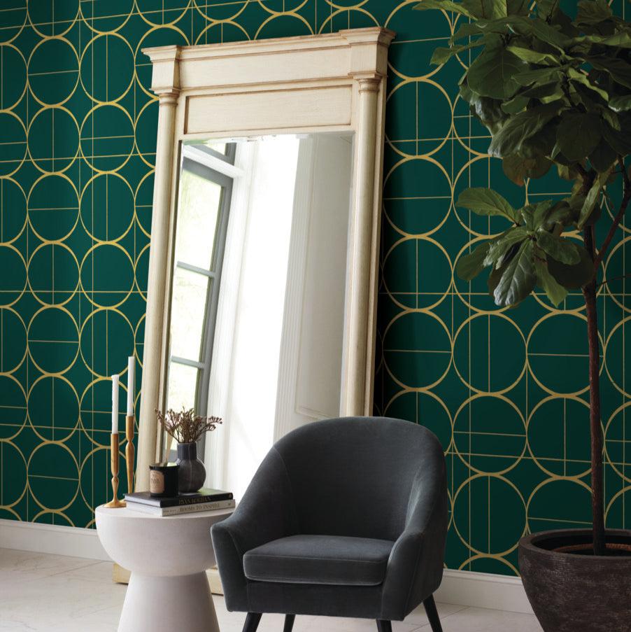 A large mirror leans against a wall covered in York Wallcoverings' Emerald & Gold Sun Circles Wallpaper. A gray chair and white side table with decor complement the stylish room, while a tall plant adds greenery to complete the look.