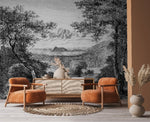 A cozy living room features two burnt orange armchairs, a woven rug, and a sideboard with a vase of dried flowers. The backdrop is Decor2Go Winnipeg's Enchanted Grove Wallpaper Mural, depicting a lush landscape with trees and mountains that creates natural tranquility.