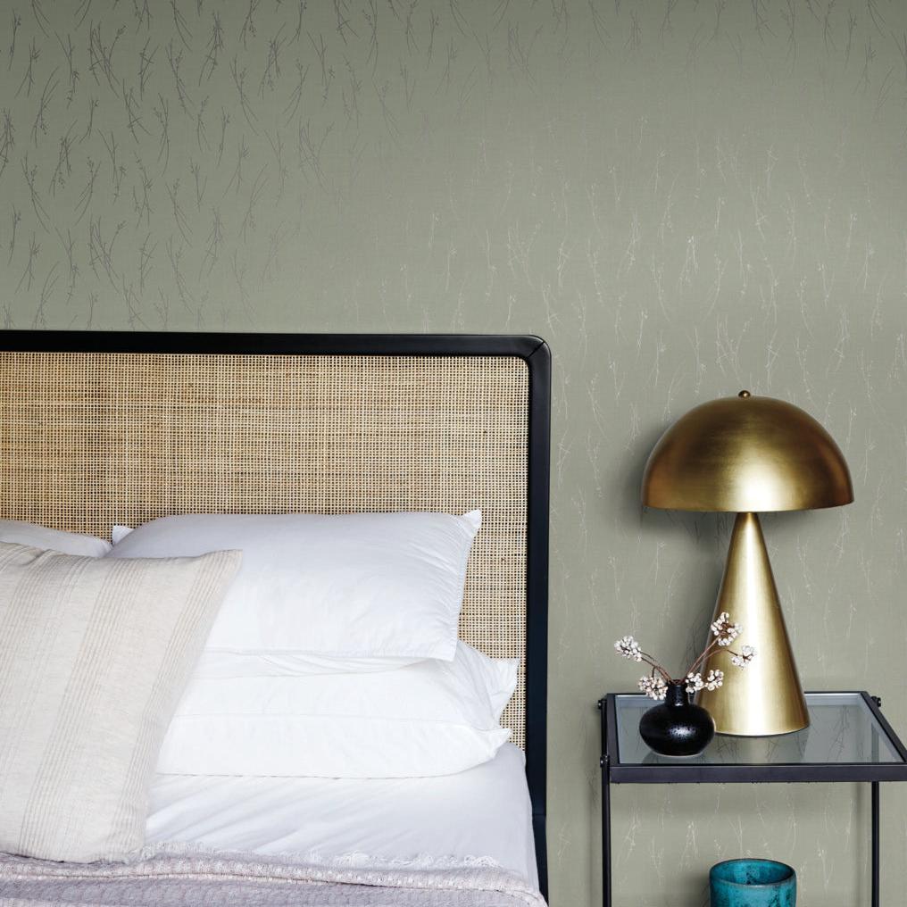 A cozy bedroom showcases a rattan headboard, white and beige pillows, and a neatly made bed. A golden dome lamp from the Modern Metals Collection sits on a black bedside table with a vase of eucalyptus and silver sprigs plus a blue candle. The wall features York Wallcoverings' Eucalyptus & Silver Sprigs Wallpaper.