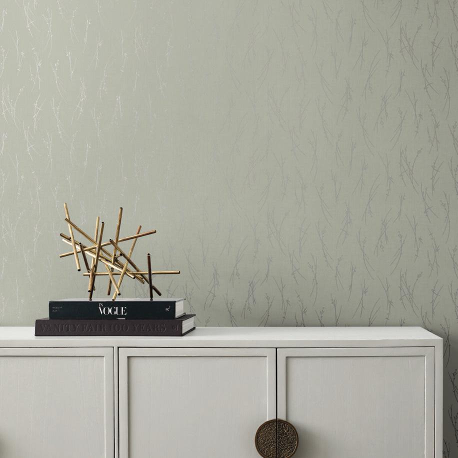 A minimalistic room showcases a light gray cabinet with round brass handles. Stacked on top are books "Vogue" and "Vanity Fair, Love," and a modern wooden sculpture from the Modern Metals Collection. The backdrop features York Wallcoverings' Eucalyptus & Silver Sprigs Wallpaper with subtle patterns.