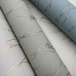 Three rolls from York Wallcoverings' Eucalyptus & Silver Sprigs Wallpaper showcase delicate branch patterns in metallic silver and gold, set against muted eucalyptus green and blue shades for a sophisticated look.