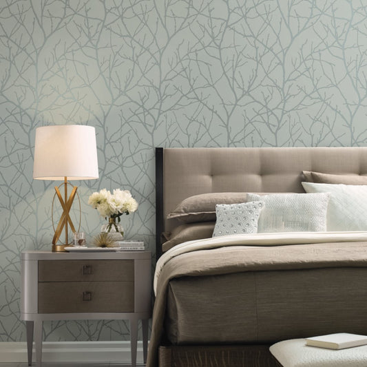 A serene bedroom boasts a cozy beige bed with pillows next to a nightstand adorned with a lamp, white flowers, and books. The walls feature York Wallcoverings' Eucalyptus & Silver Trees Silhouette Wallpaper, enhancing the room's cozy elegance.