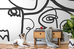 A stylish room featuring a large Face Art Wallpaper Mural from Decor2Go Wallpaper Mural on the wall, a wooden console table with a red mug atop, a draped grey blanket, and a plant. A white and brown dog lounges on