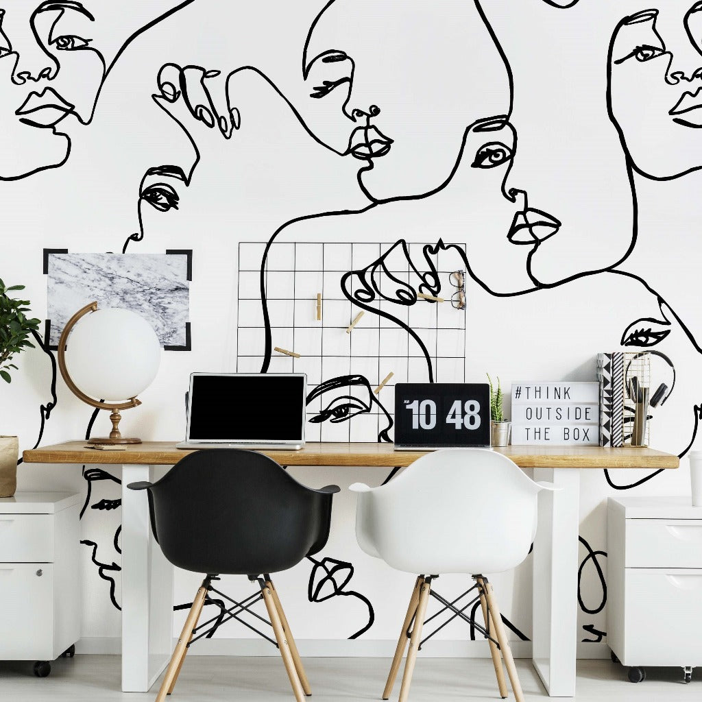 A modern home office with a desk featuring a laptop, a globe, and decorative items. The wall behind is adorned with a unique Familiar Faces Wallpaper Mural from Decor2Go Wallpaper Mural depicting abstract faces. Two chairs are in front.