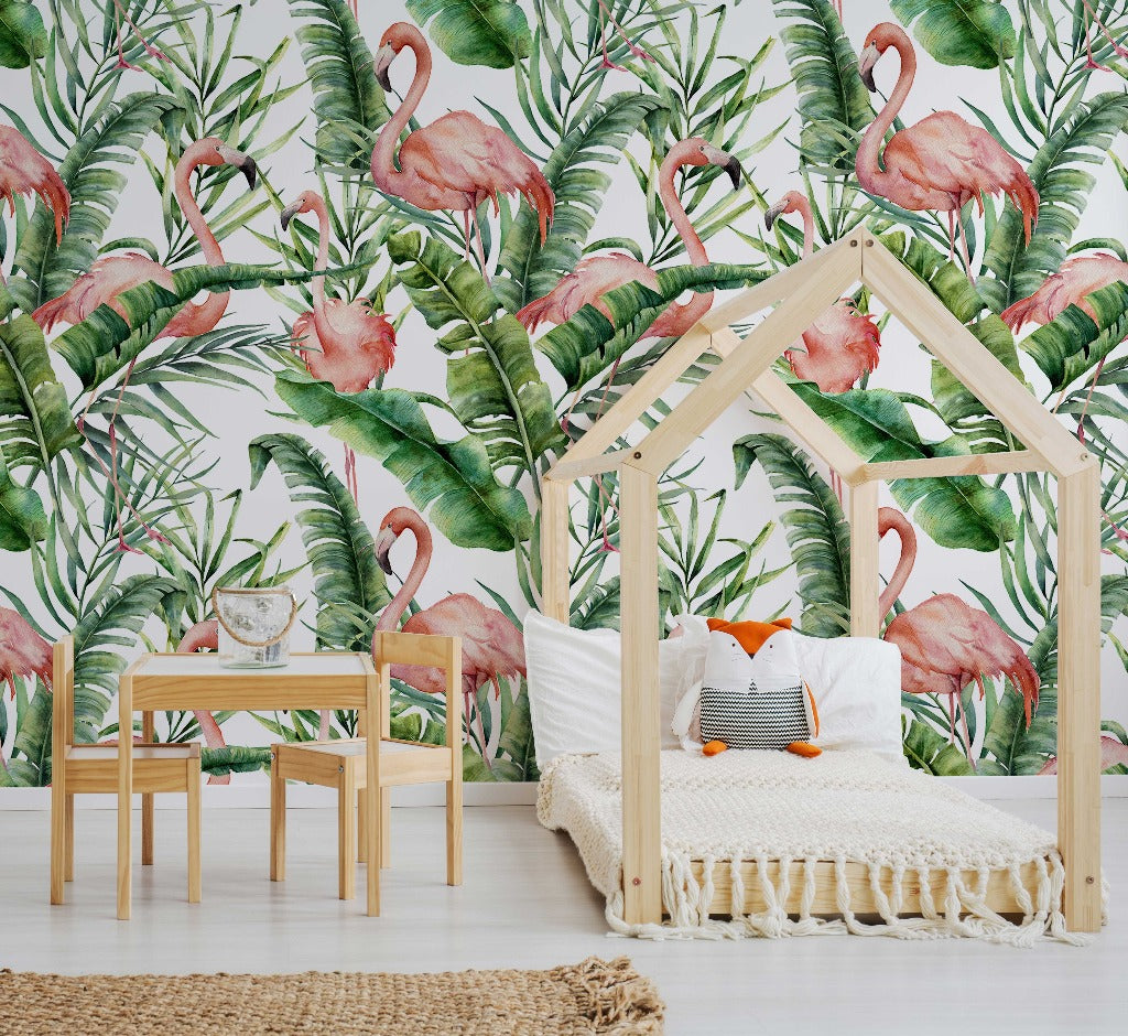 A stylish children's room featuring a house-shaped bed frame with a white mattress, small wooden table and chairs, Decor2Go Wallpaper Mural Flamingos and Tropical Leaves wallpaper mural with lush green foliage, and a woven rug on the floor.