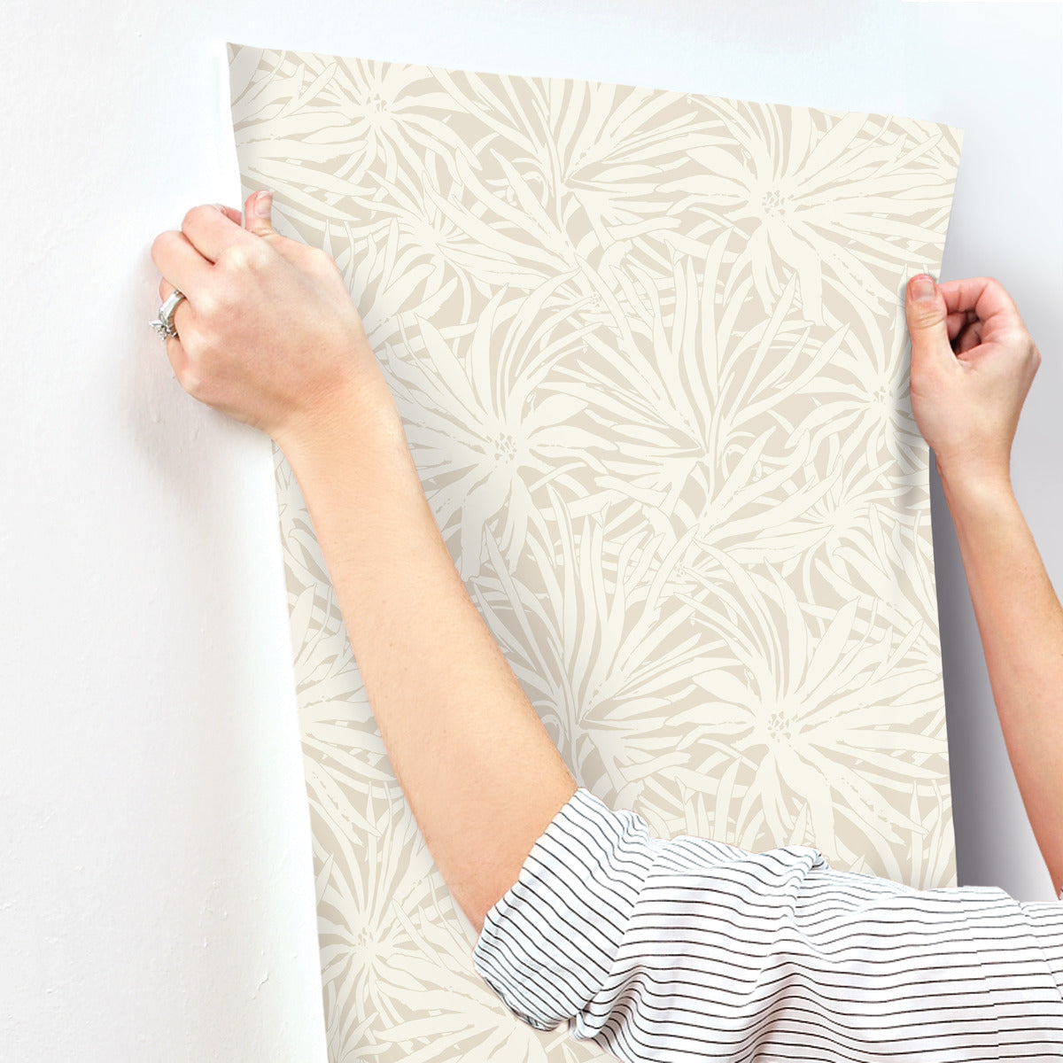 A person in a striped shirt uses both hands to align York Wallcoverings Floral Canopy Linen Wallpaper (60 SqFt), featuring a subtle beige floral design, as they smooth it onto a white wall.