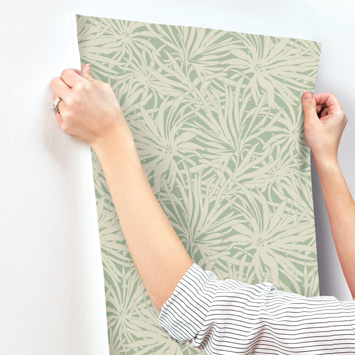 A person with rolled-up sleeves is using both hands to smooth York Wallcoverings' Floral Canopy Olive Wallpaper (60 SqFt), featuring a beige leafy pattern, onto a white wall.