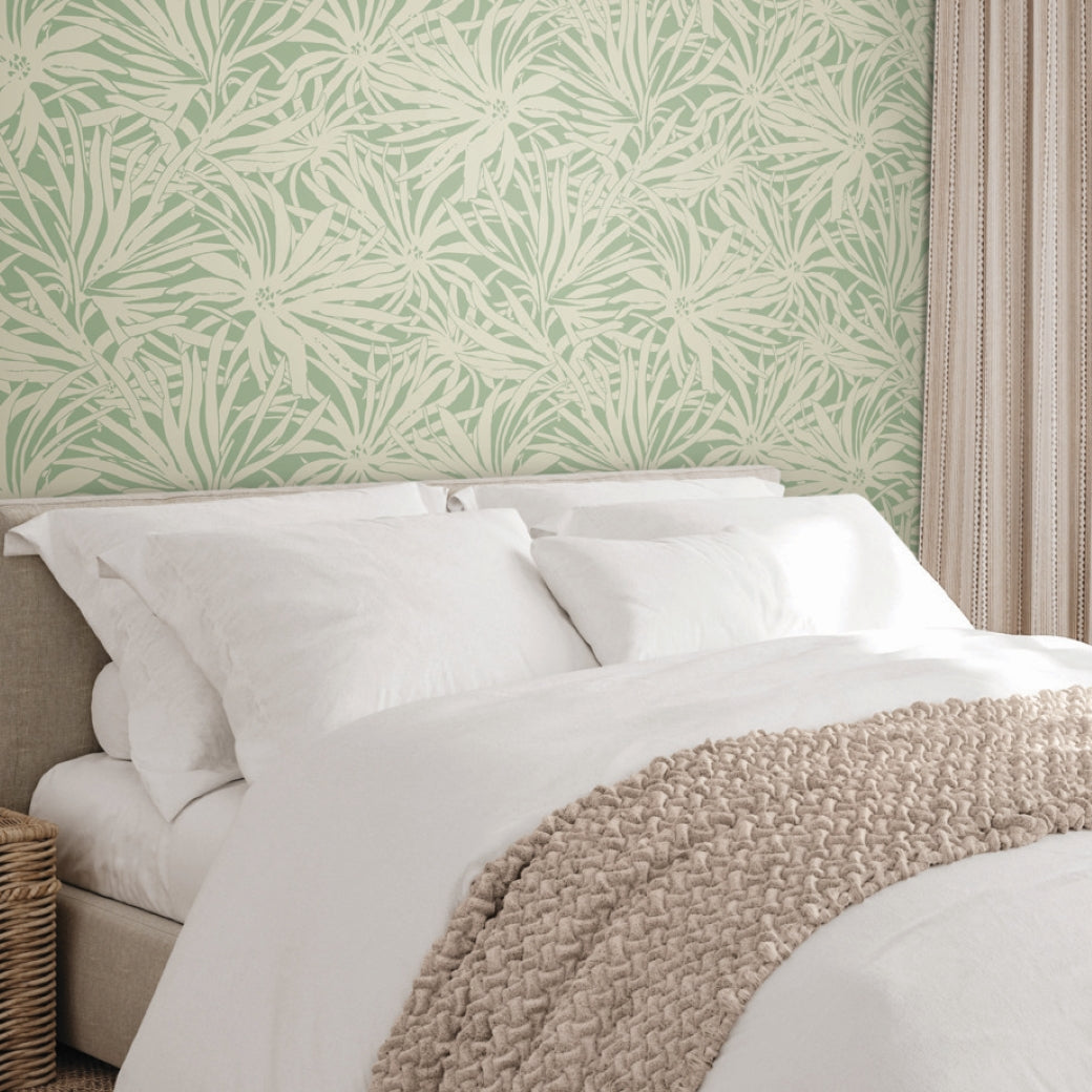 A neatly made bed with white sheets and pillows, a beige knit blanket, and York Wallcoverings’ Floral Canopy Olive Wallpaper (60 SqFt) featuring a botanical pattern; a wicker basket sits beside the bed.