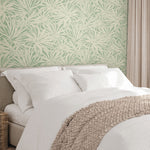 A neatly made bed with white sheets and pillows, a beige knit blanket, and York Wallcoverings’ Floral Canopy Olive Wallpaper (60 SqFt) featuring a botanical pattern; a wicker basket sits beside the bed.