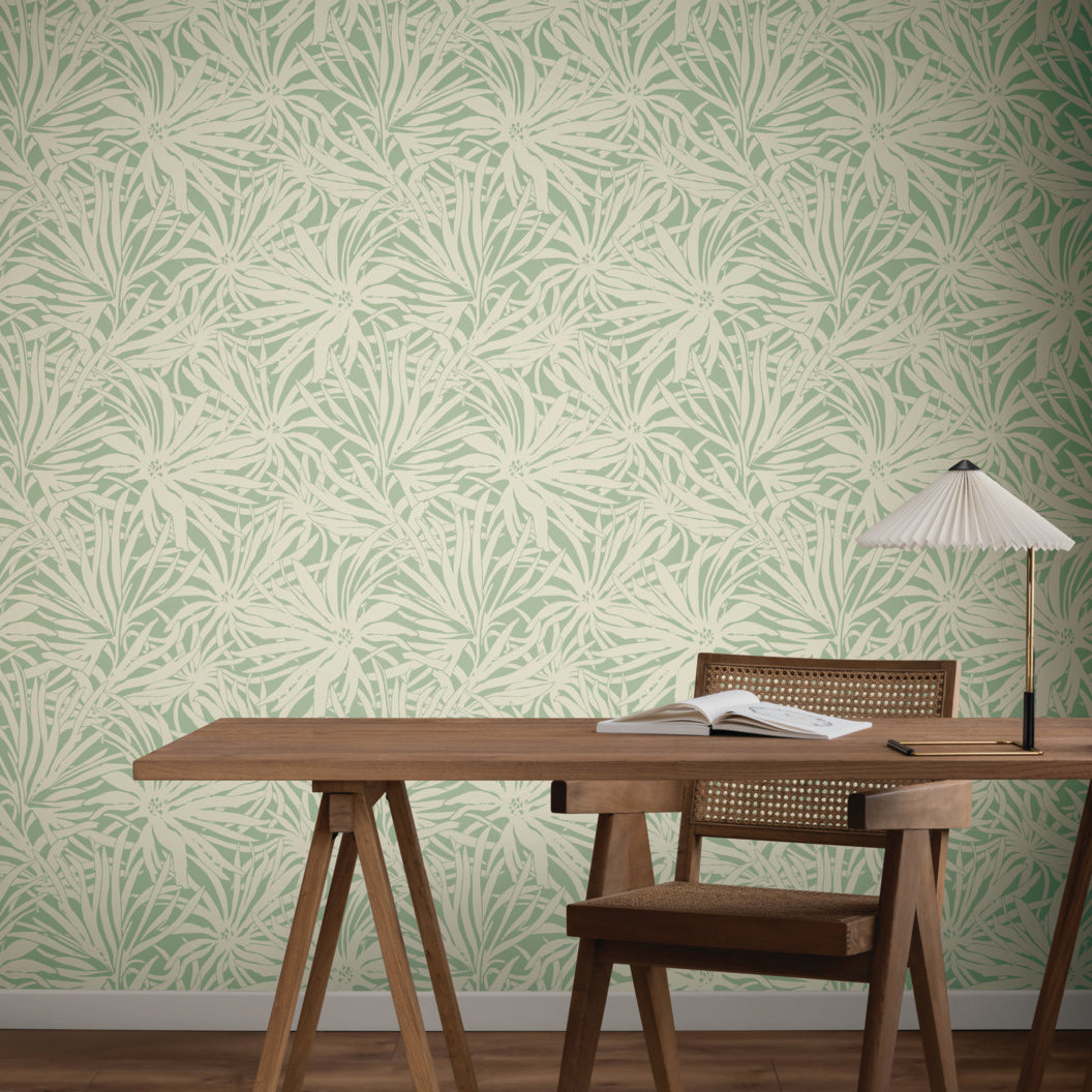 A minimalist workspace features a wooden desk, chair, lamp, open notebook, and York Wallcoverings' Floral Canopy Olive Wallpaper (60 SqFt), adding calming green and cream botanical patterns to the background.