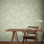 A minimalist workspace features a wooden desk, chair, lamp, open notebook, and York Wallcoverings' Floral Canopy Olive Wallpaper (60 SqFt), adding calming green and cream botanical patterns to the background.