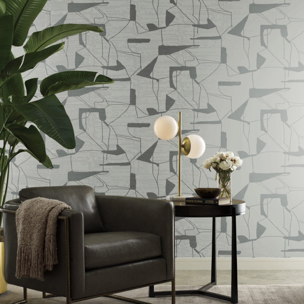 A modern living room with a gray armchair, a side table holding a double-bulb lamp, and a vase of white flowers. The backdrop is Fog & Silver Abstract Geo Wallpaper by York Wallcoverings from the Modern Metals Collection in abstract patterns, complemented by a large potted plant.